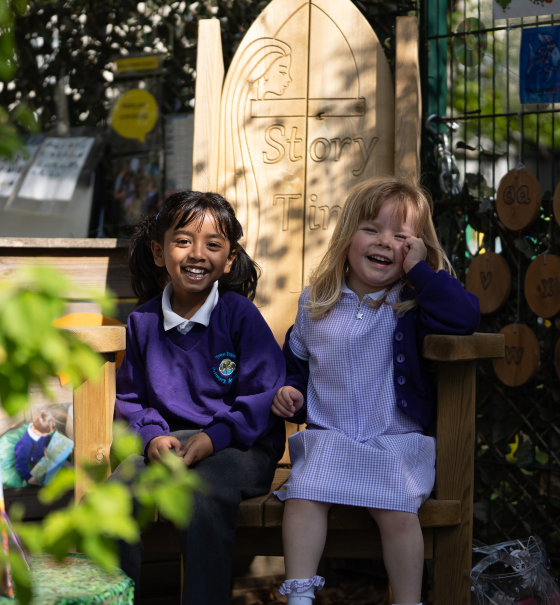 Two young girls are seen sat together on a large wooden chair outdoors on the school grounds. They each have a bright smile on their faces.