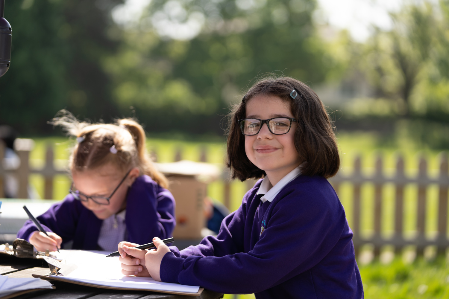 Two young girls are pictured sitting outdoors on the school grounds, writing on sheets of paper attached to clipboards. One of the girls is seen looking up and smiling at the camera.