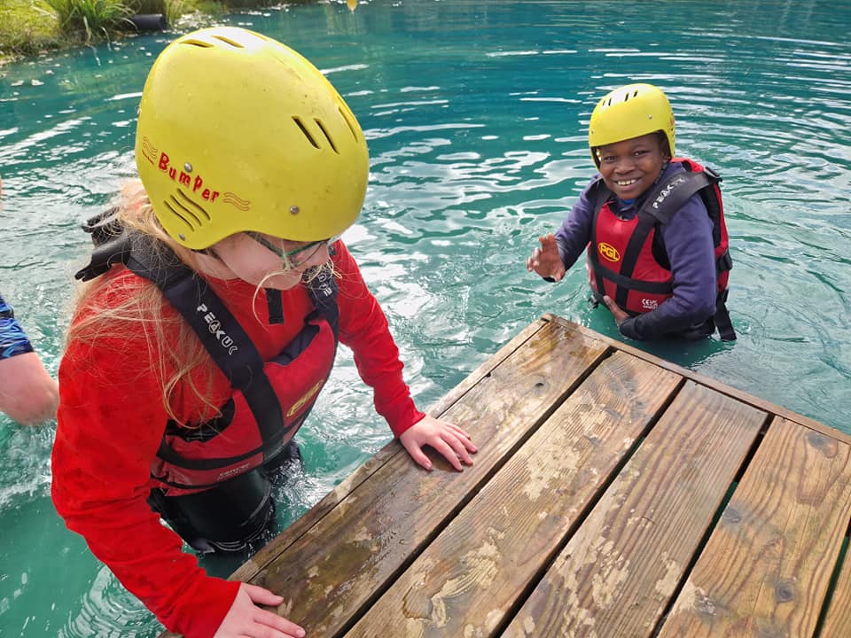 2 students in life jackets and helmets smiling while climbing out of the water