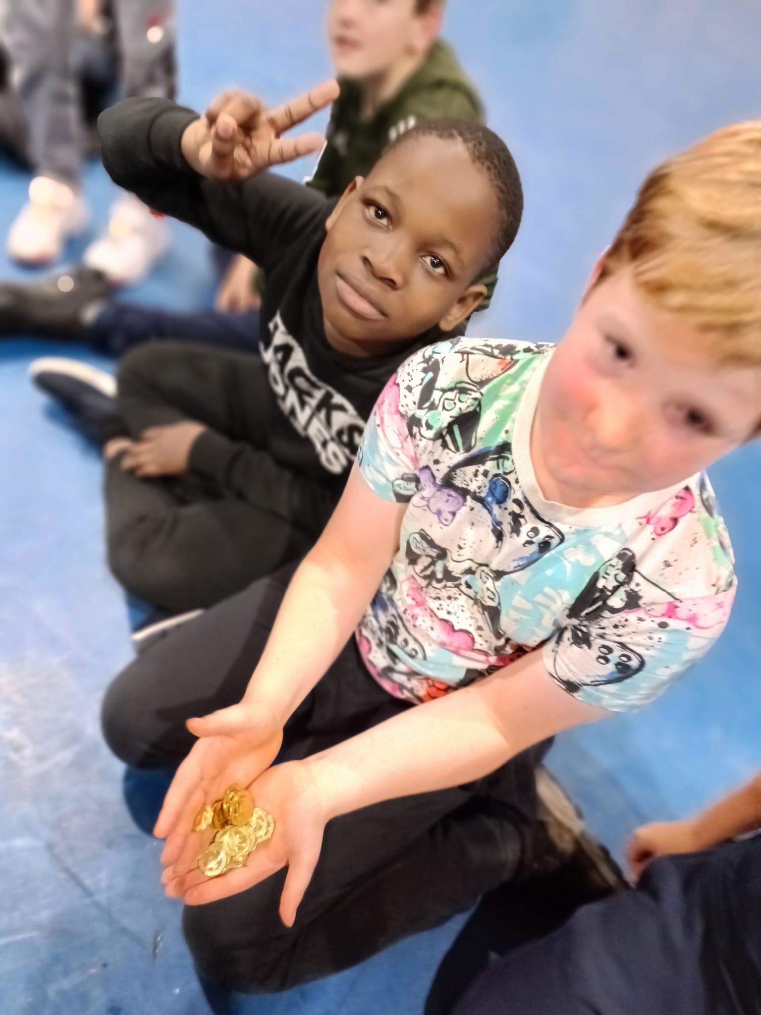 2 children sitting on the floor of a sports hall, one holding out a handful of gold coins