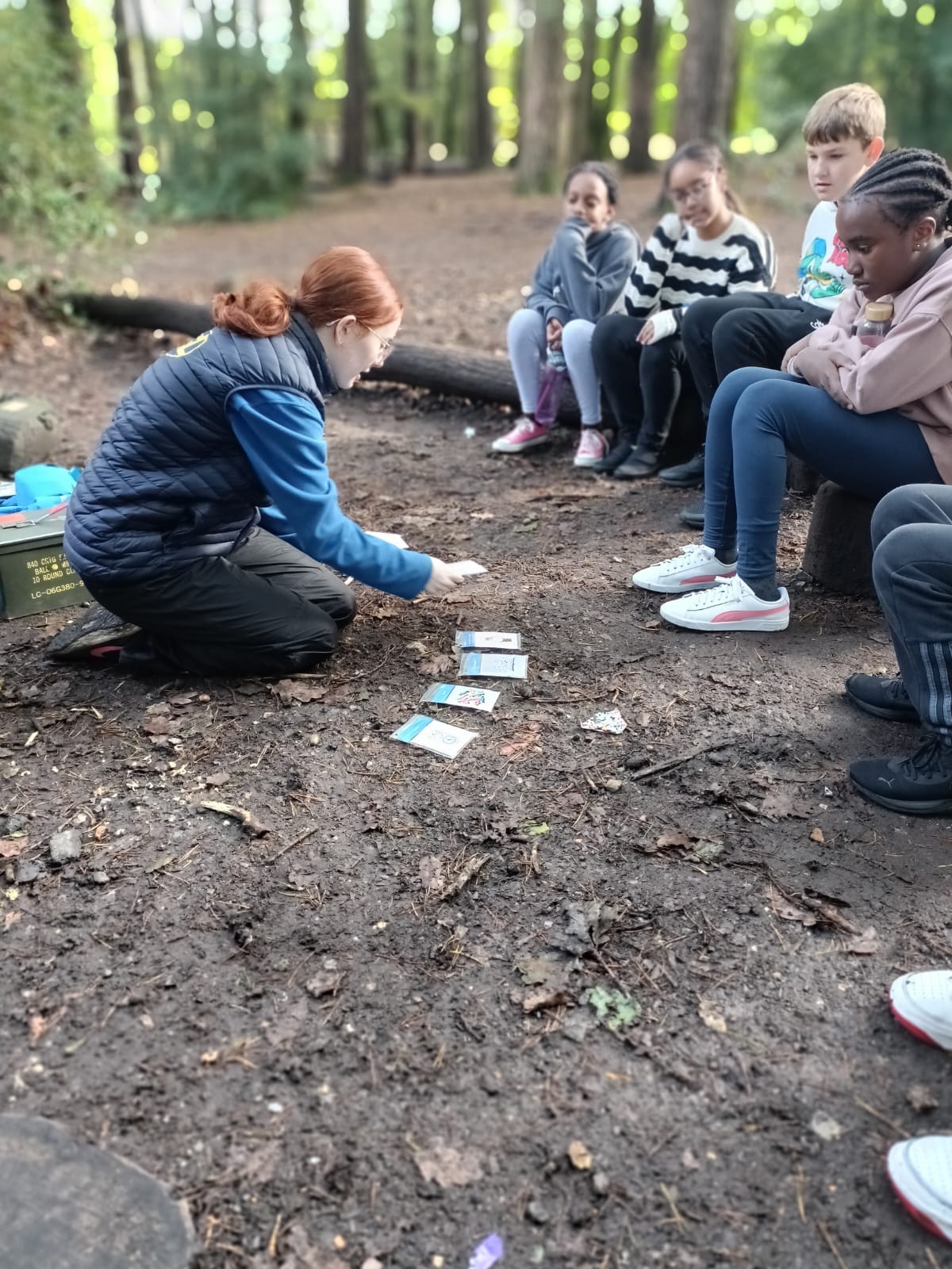 An instructor laying plastic cards on the floor while a group of children sit on a log watching