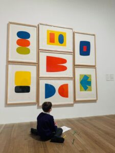 A student sat on the floor drawing, in front of a piece of artwork on the wall